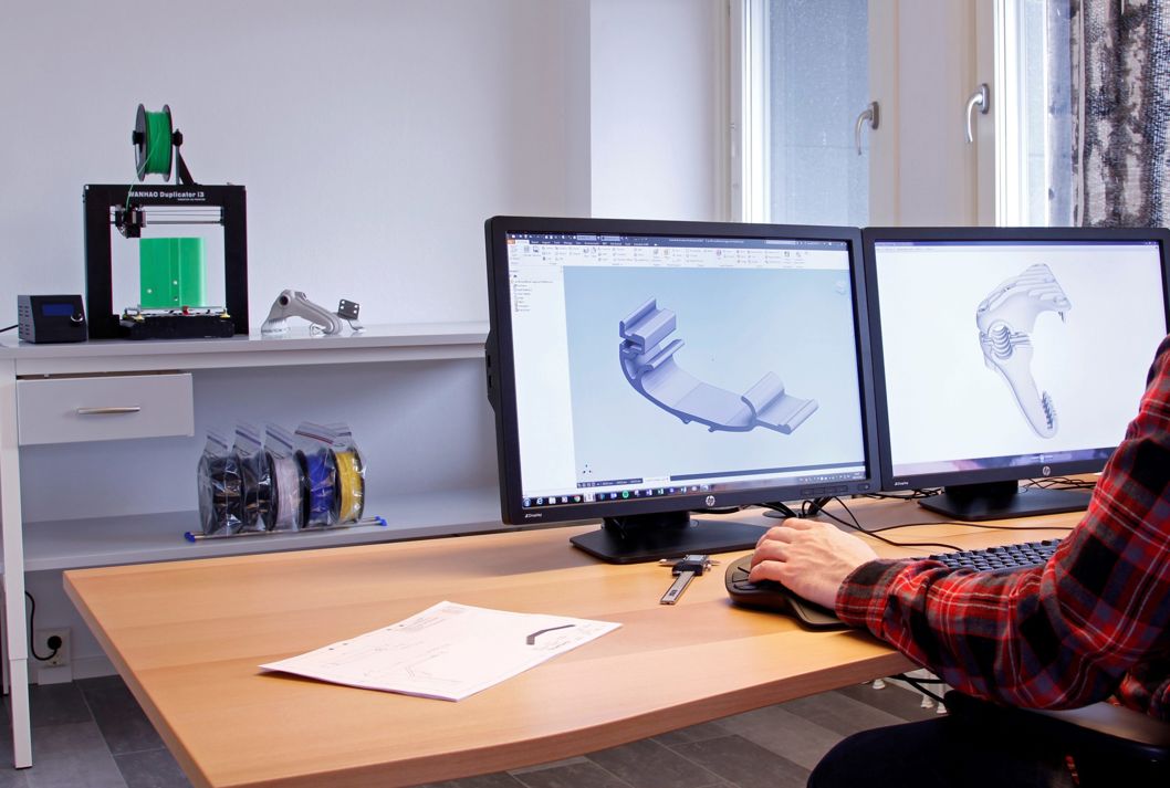 Person sitter vid skrivbord och arbetar med 3D-modell av bryggtillbehör på skärm, 3D-printer i bakgrunden, Floattechs kontor