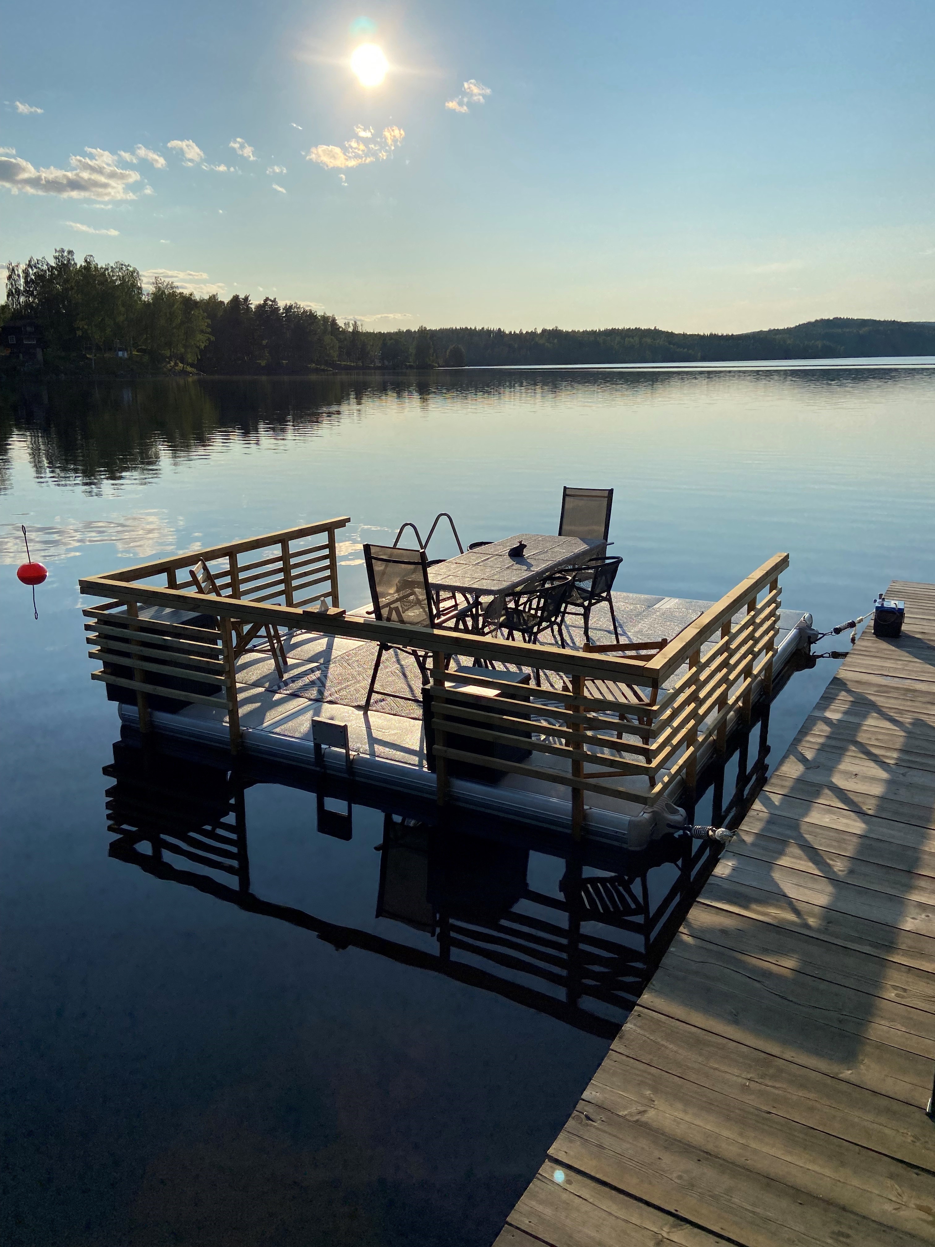 EasyFloat brygga 3,6 × 4,2 m i Värmland med egenbyggda trä-räcken, badstege, solstolar, bord och motorfäste på spegelblankt vatten en sommardag
