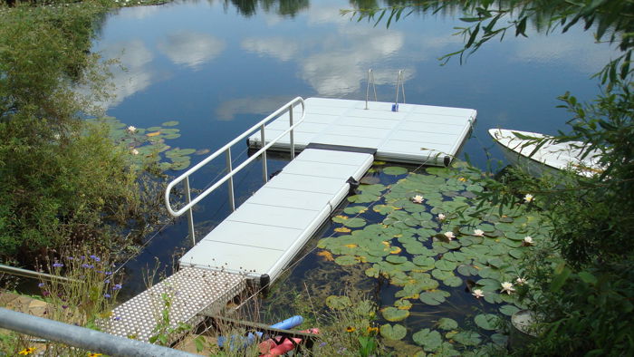 EasyFloat brygga i Tyskland med landgång, räcke och badstege. En båt ligger intill bryggan och näckrosor flyter på det lugna vattnet.