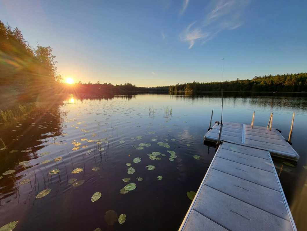 Privat EasyFloat-brygga med badstege på stilla vatten vid solnedgång. Näckrosor flyter nära bryggan och ett fiskespö lutar mot bryggans kant.