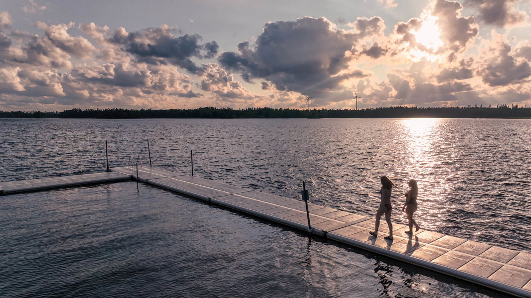 Stor offentlig EasyFloat flytbrygga vid en badplats med öppet vatten och dramatiska moln i solnedgång, två personer promenerar på bryggan.